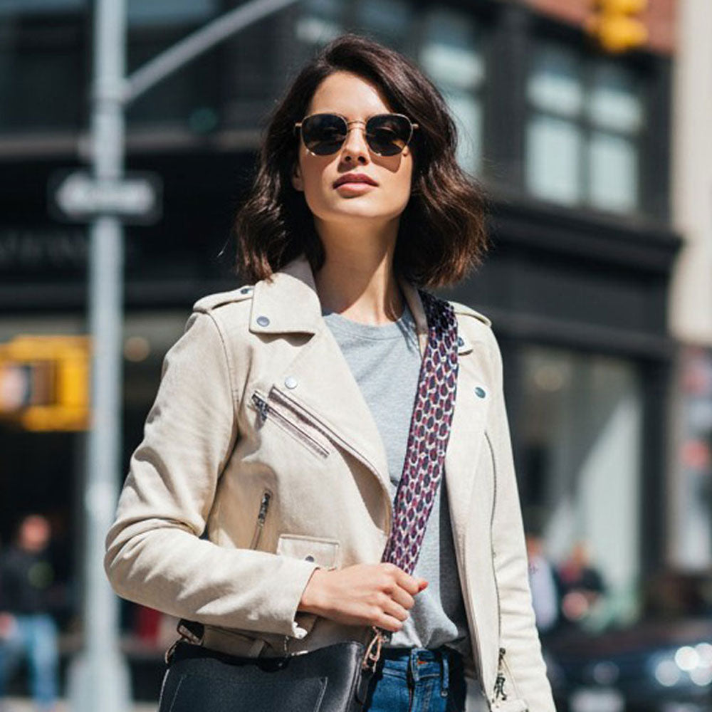 Woman wearing sunglasses and a beige jacket on a city street. Wearing a pattern bag strap and black crossbody bag.