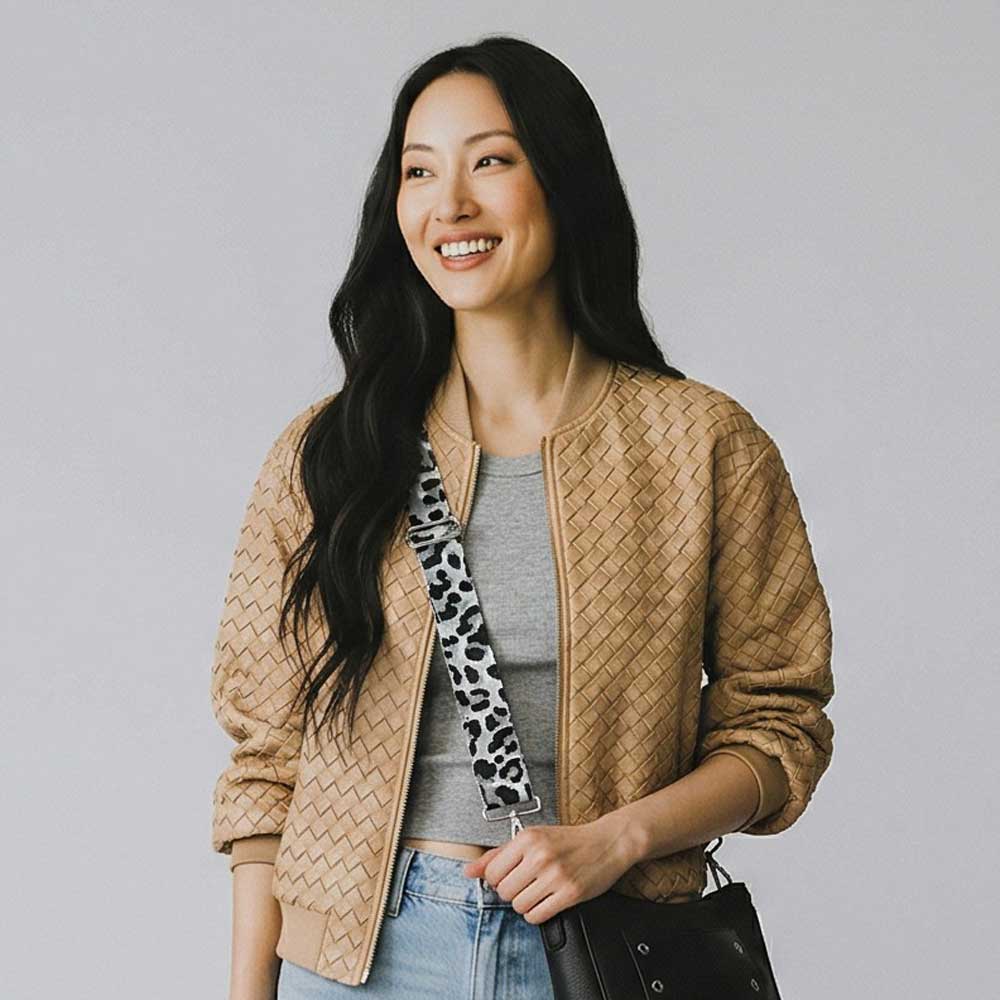 Woman wearing a beige quilted jacket with a gray top and blue jeans, wearing a black handbag with a grey leopard bag strap against a plain background.
