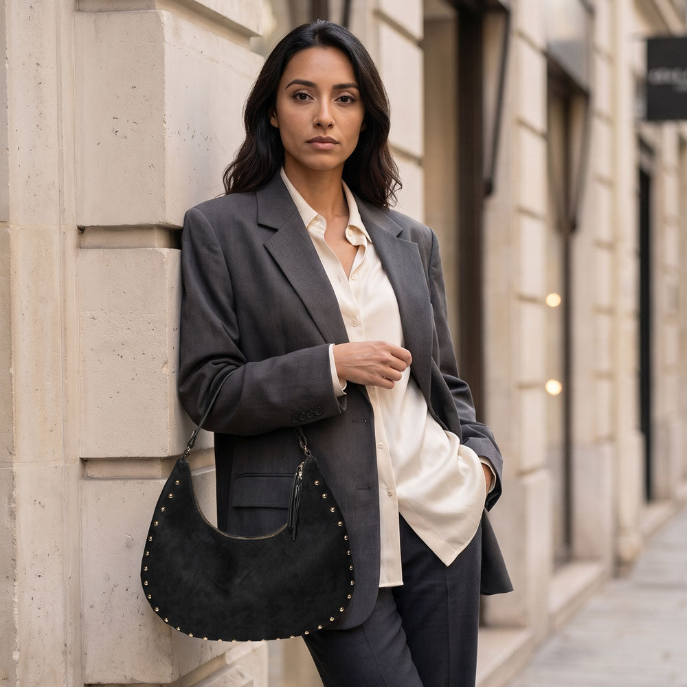 A woman stands on a city sidewalk against a beige stone building, wearing a dark gray blazer over a cream button-up shirt and dark trousers, while carrying a black crescent-shaped suede handbag with gold-tone studs.