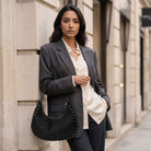 A woman stands on a city sidewalk against a beige stone building, wearing a dark gray blazer over a cream button-up shirt and dark trousers, while carrying a black crescent-shaped suede handbag with gold-tone studs.