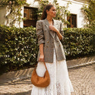 Woman in a plaid blazer and white dress holding a camel vegan suede with studs handbag on a street.