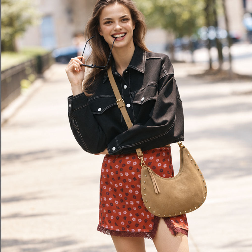 A woman stands on a sunlit city sidewalk wearing a black denim jacket, a red floral skirt, and carrying a tan suede crescent shoulder bag with silver studs and a tan strap.