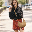 A woman stands on a sunlit city sidewalk wearing a black denim jacket, a red floral skirt, and carrying a tan suede crescent shoulder bag with silver studs and a tan strap.