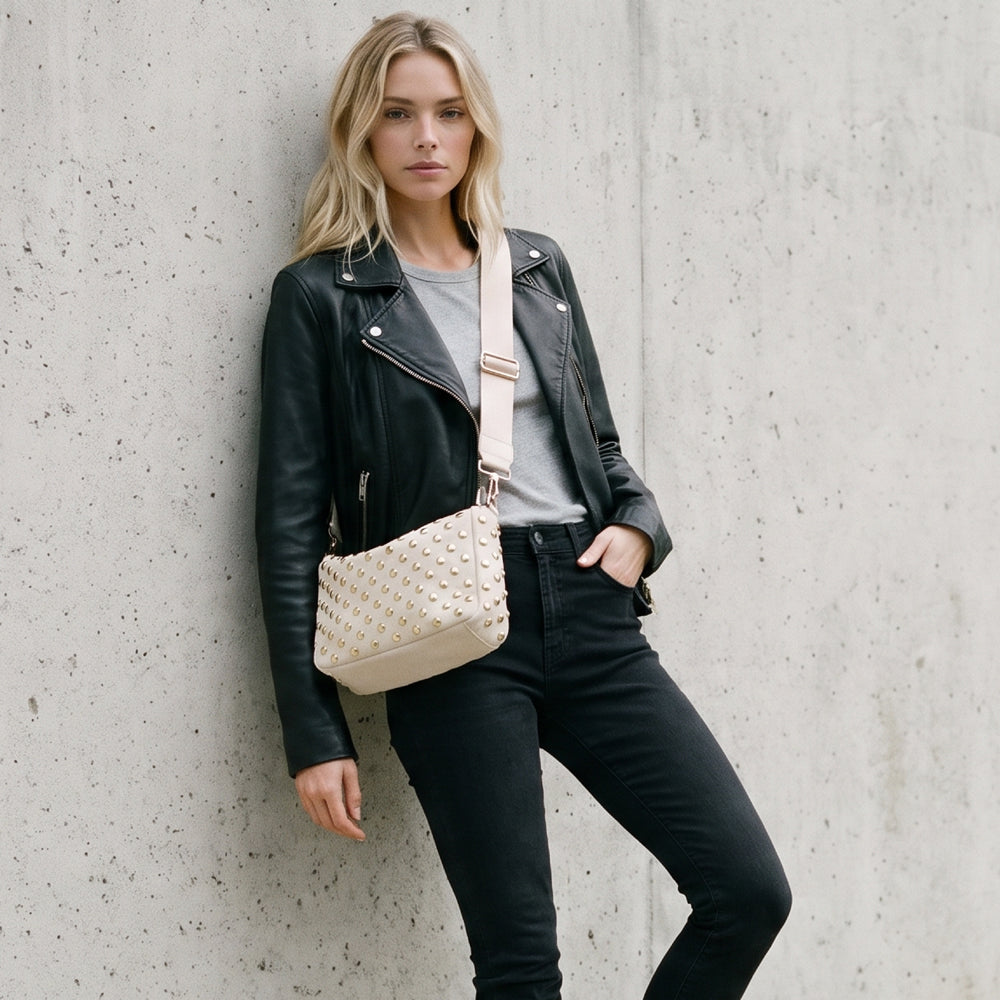 A blonde woman posing against a textured concrete wall, wearing a black leather moto jacket, black jeans, and a gray t-shirt, while carrying a cream vegan suede crossbody bag with a studded pattern and solid bag strap.