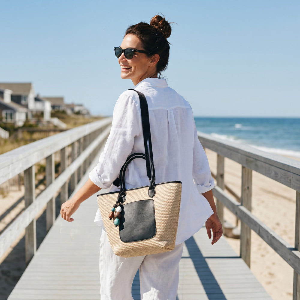 Woman walking on a beach boardwalk with a raffia tote, wearing sunglasses and a white outfit.