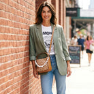 Woman leaning against a brick wall, wearing a green blazer and blue jeans, with a deer print handbag.