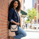 Woman leaning against a brick wall on a city street, wearing a navy blazer and blue jeans with a handbag.