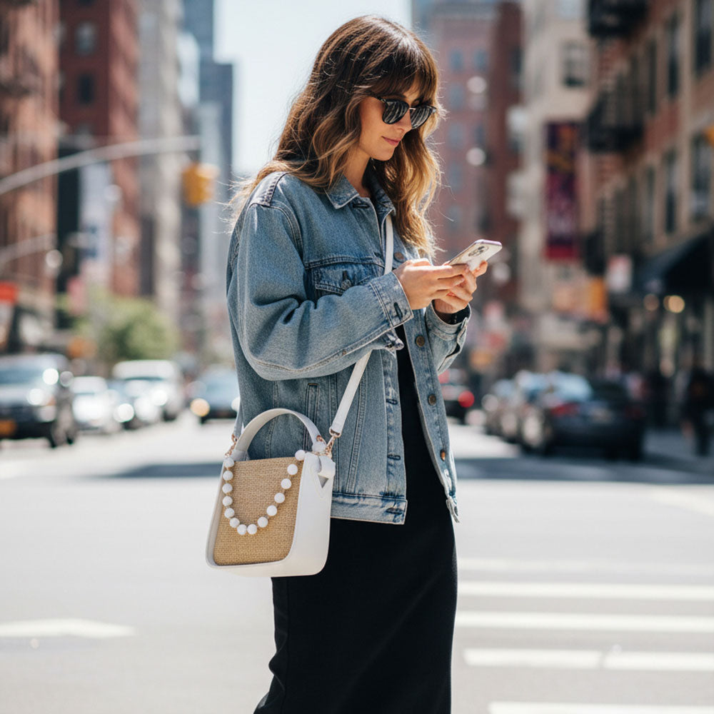 Woman in a denim jacket using a phone on a city street.  Wearing a raffia handbag.
