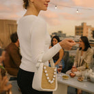 Woman holding a white handbag with a raffia design at an outdoor event.