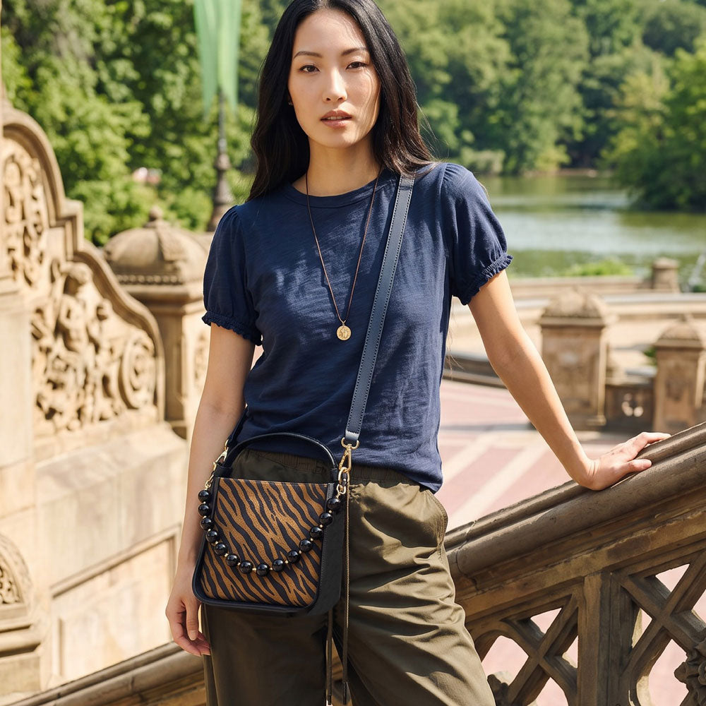 A woman stands on an outdoor stone bridge wearing a navy blue short-sleeve t-shirt, olive green pants, and a crossbody bag with a dark brown and black tiger-stripe pattern and a beaded strap.
