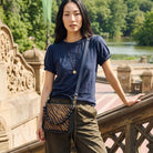 A woman stands on an outdoor stone bridge wearing a navy blue short-sleeve t-shirt, olive green pants, and a crossbody bag with a dark brown and black tiger-stripe pattern and a beaded strap.