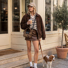 A woman stands on a city sidewalk in front of a beige building, wearing an oversized brown suede jacket, a brown mini skirt, a striped shirt, white socks, and a beige cap. She carries a leopard-print crossbody bag and holds a coffee cup while walking a small brown and white dog.