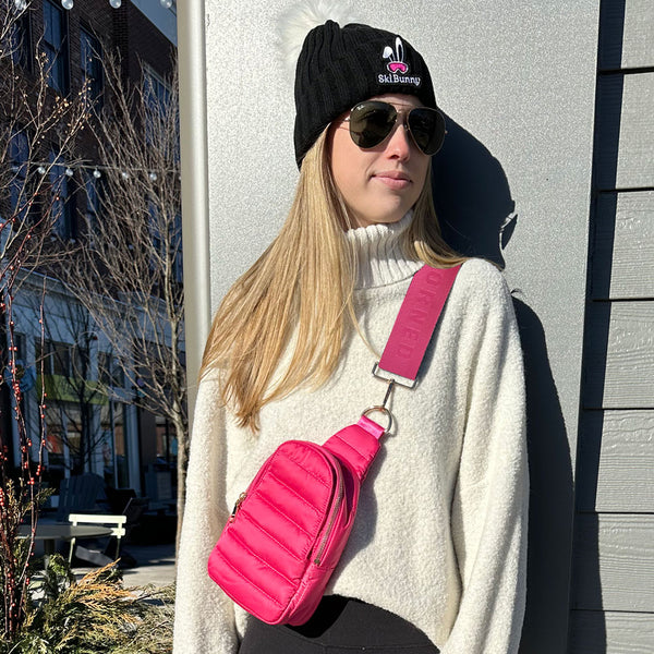Person wearing a black beanie with a pink bunny design, sunglasses, and a white sweater with a pink crossbody bag.