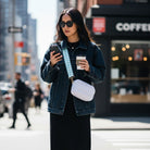 Woman in a city street holding a phone and a coffee cup, wearing a denim jacket and sunglasses.  And wearing a white crossbody bag.