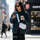 Woman in a city street holding a phone and a coffee cup, wearing a denim jacket and sunglasses.  And wearing a white crossbody bag.