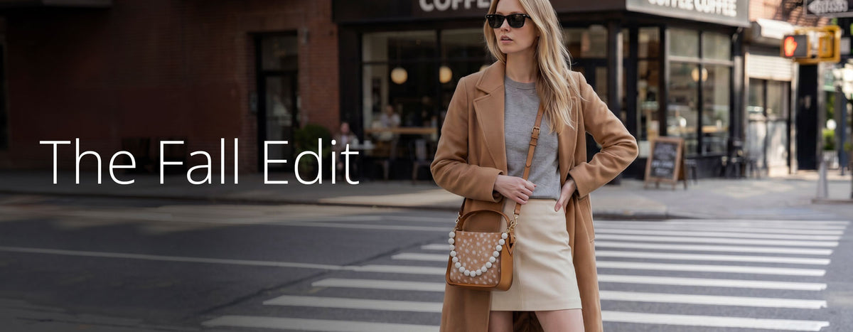 A woman stands on a city crosswalk wearing a tan coat, light gray top, cream skirt, and a brown crossbody bag with pearl detailing. The background shows a blurred street scene with storefronts.