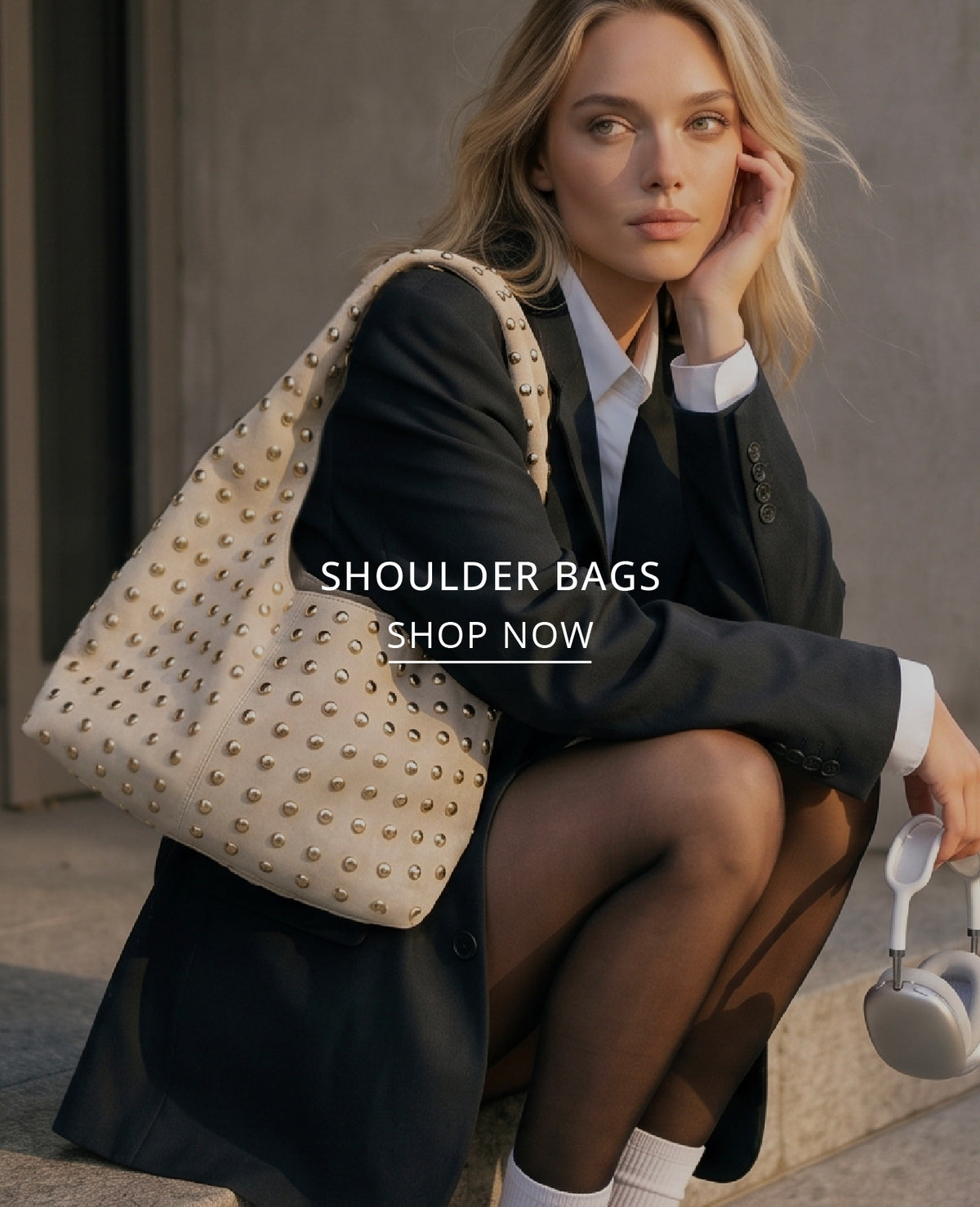A woman in a black blazer and white shirt sits on stone steps, wearing a large cream shoulder bag covered in gold studs. The text 'SHOULDER BAGS SHOP NOW' is overlaid on the center of the image.