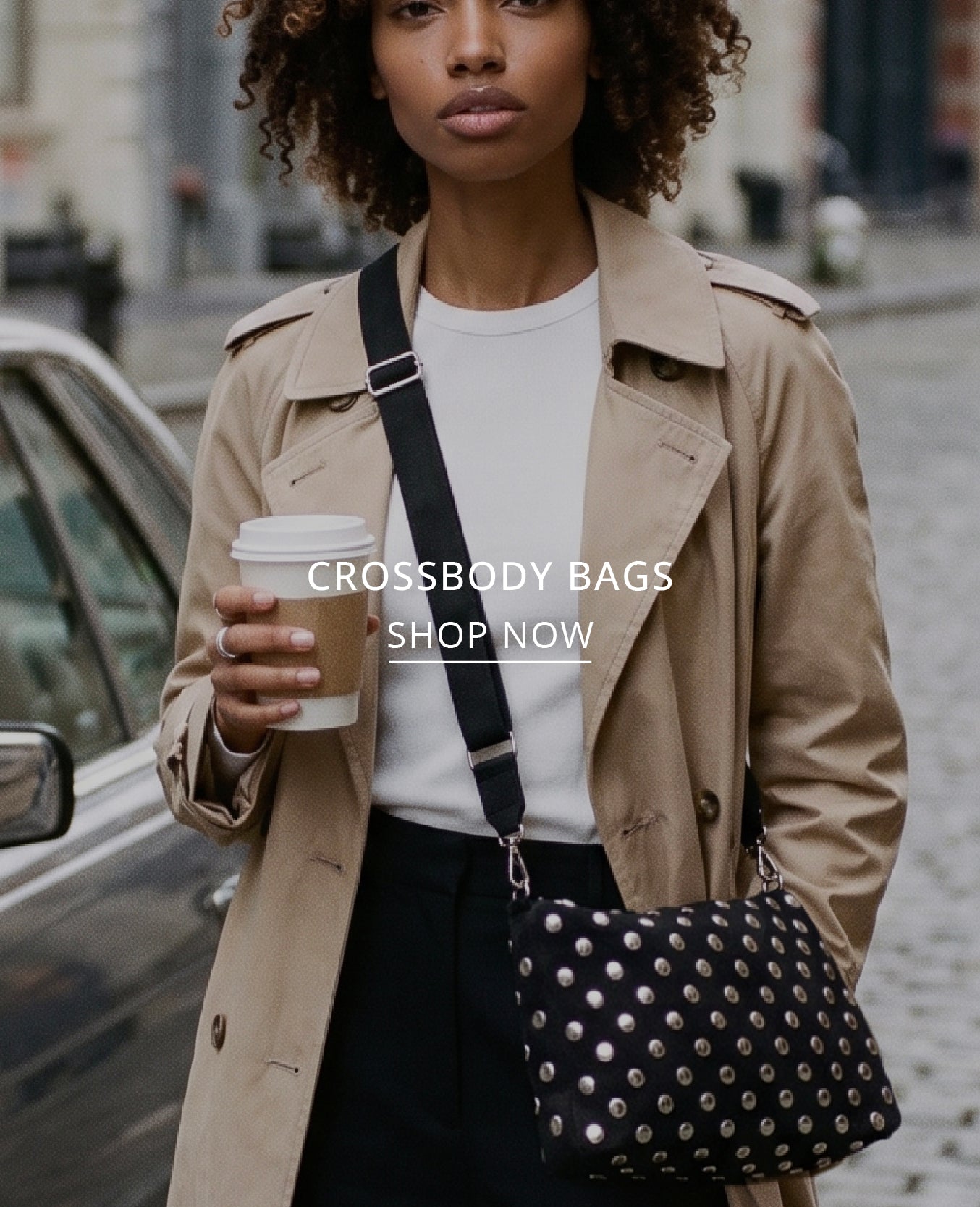 A woman wearing a beige trench coat and white top carries a black crossbody bag with silver studs and a black strap, holding a coffee cup on a city street. White text overlay reads 'CROSSBODY BAGS SHOP NOW'.