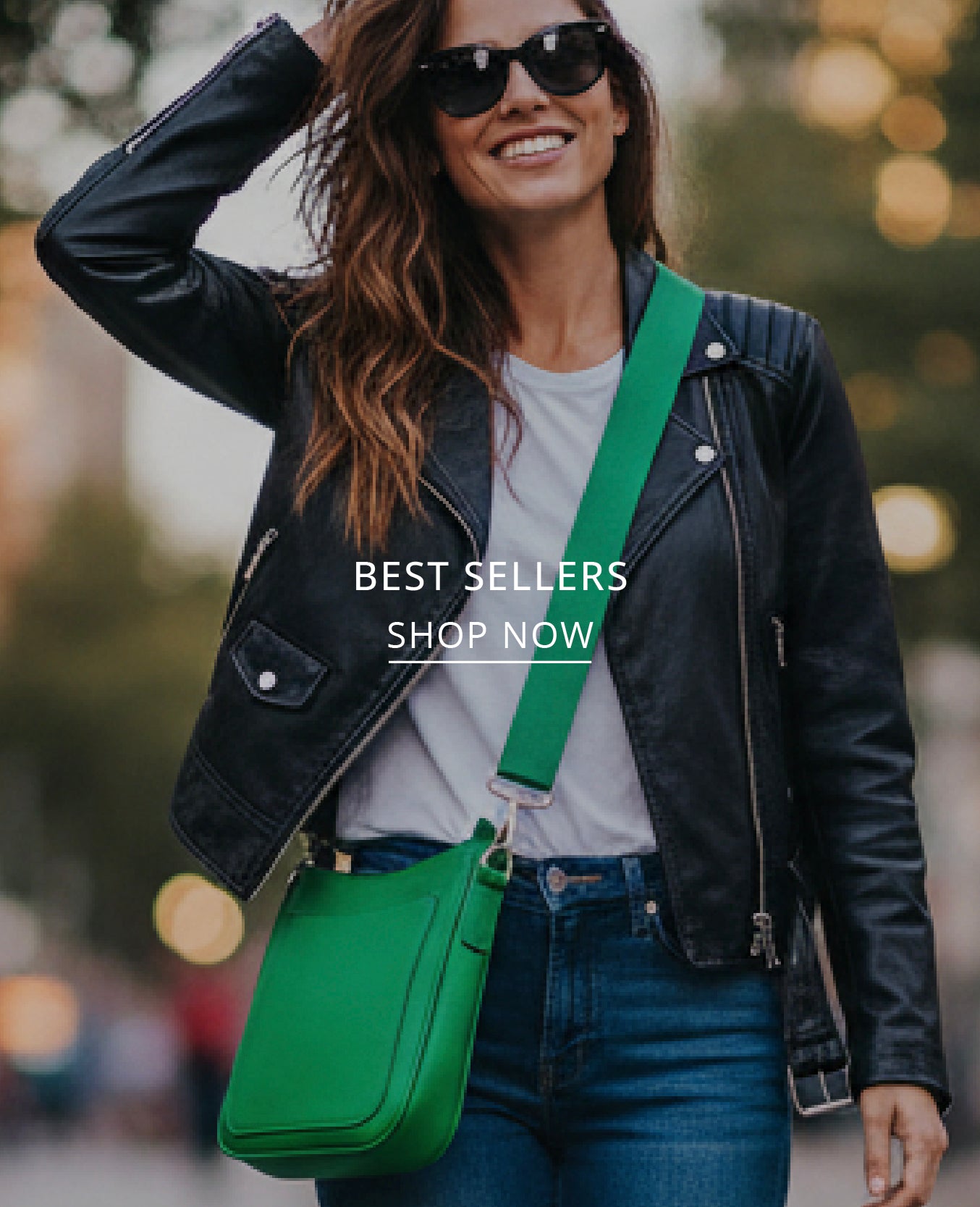 A woman wearing a black leather jacket, white t-shirt, and blue jeans carries a bright green crossbody bag with a wide strap. She is wearing black sunglasses and smiling, with a blurred outdoor background and the text 'BEST SELLERS SHOP NOW' overlaid in the center.