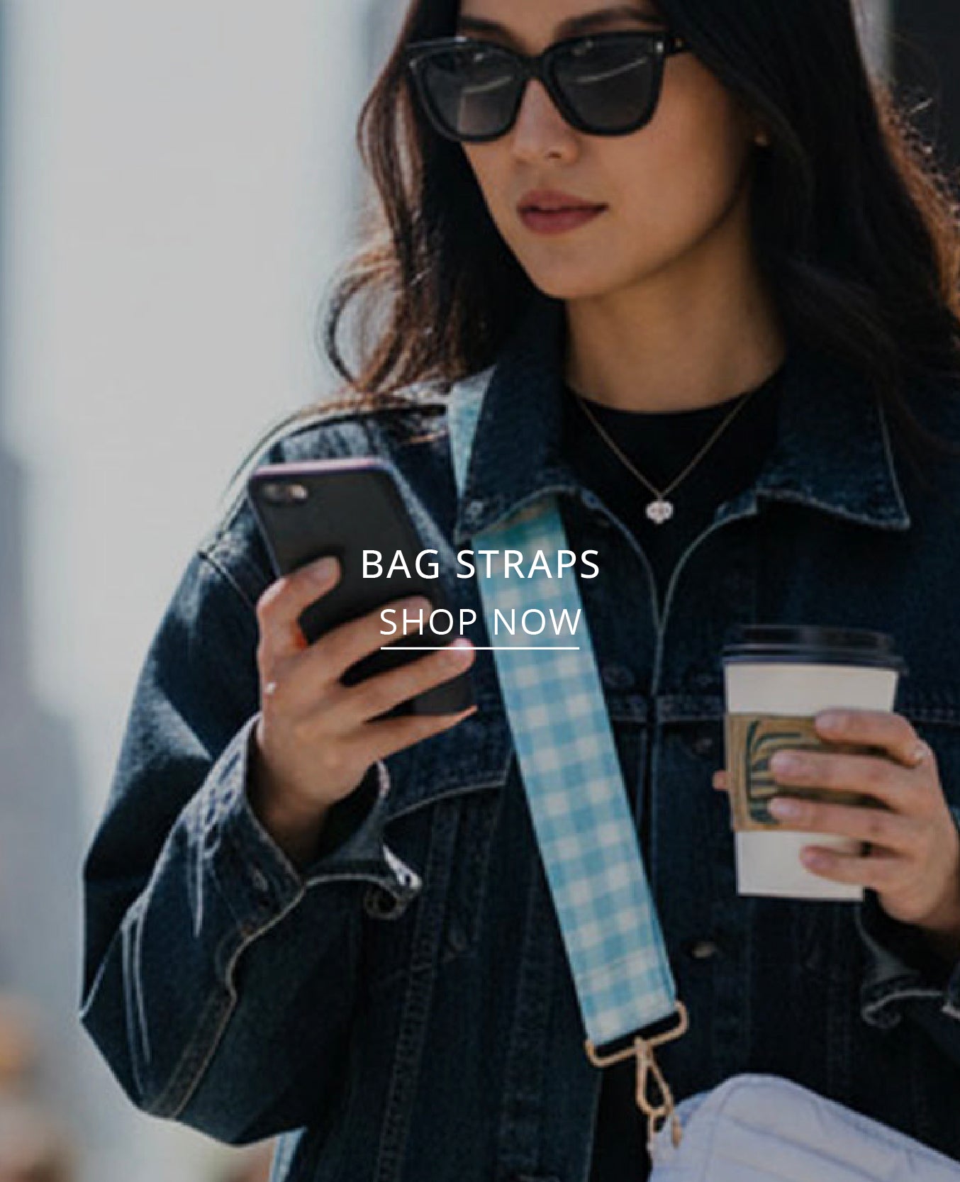 A woman wearing black sunglasses, a dark denim jacket, and holding a coffee cup and phone, with a light blue and white gingham bag strap diagonally across her body. White text overlay reads 'BAG STRAPS SHOP NOW'.