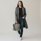 Woman holding a army and ecru woven neoprene tote, wearing a long coat and jeans, on a plain background