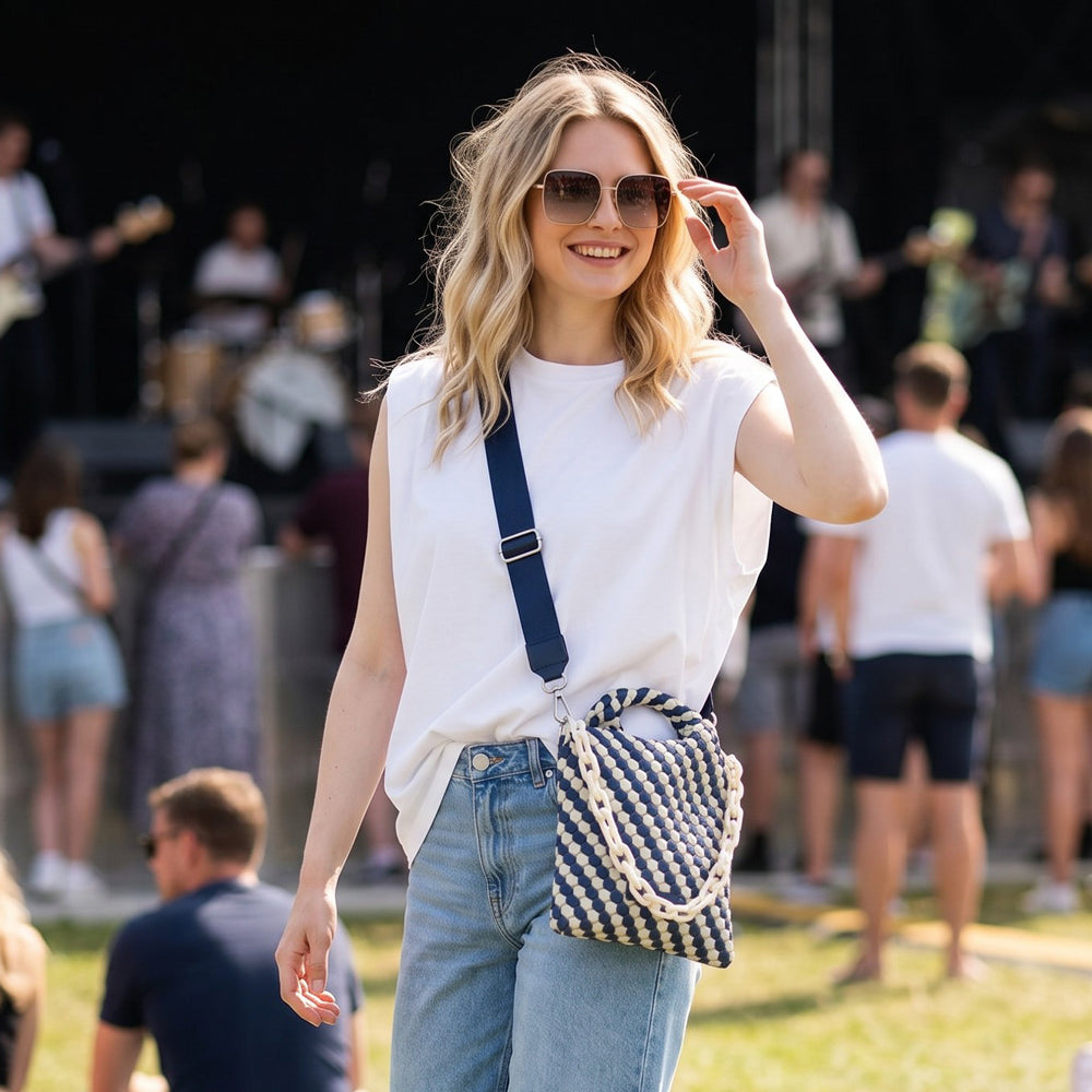 Woman in a white sleeveless top and blue jeans with a striped bag, standing outdoors with blurred people in the background.