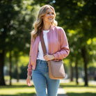 Woman in a pink jacket and blue jeans walking outdoors with a beige bag.