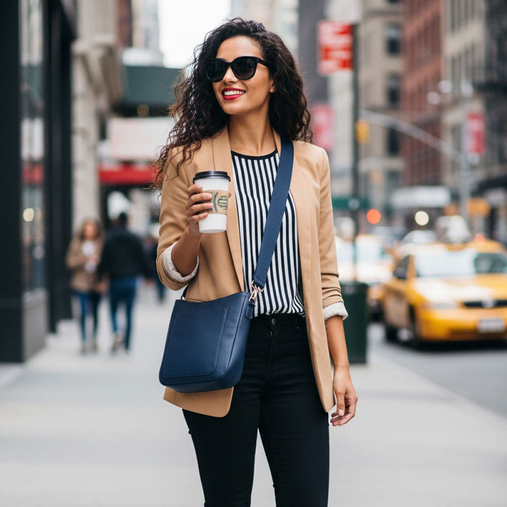 Woman walking on a city street holding a coffee cup, with a blurred background of urban scenery. She is wearing a navy crossbody bag. 