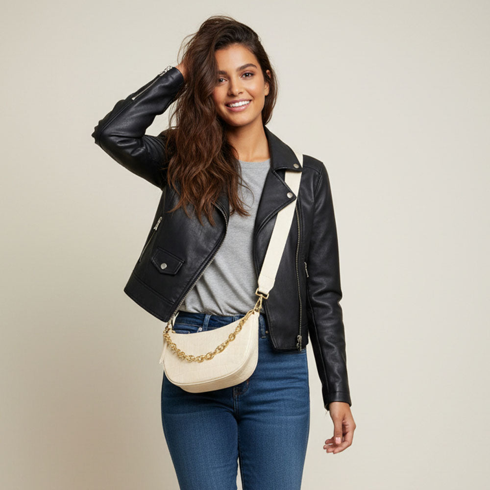 Woman wearing a black moto jacket, grey tee, denim, and a cream crossbody bag with chain detail.