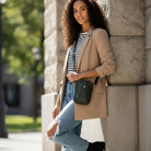 Woman in a beige coat and striped shirt leaning against a stone wall outdoors.  She is wearing an army crossbody body. 