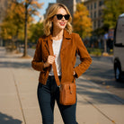 Woman in a brown jacket and sunglasses walking on a sidewalk with trees and buildings in the background wearing a camel crossbody bag