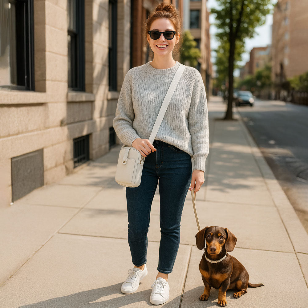 Woman walking a small dog on a city street. Wearing a cream crossbody bag.