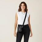 Woman wearing a white sleeveless top and black pants with a black crossbody with silver studs bag on a beige background
