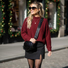 Woman wearing a red sweater and black skirt with a black bag, standing in front of Christmas decorations.