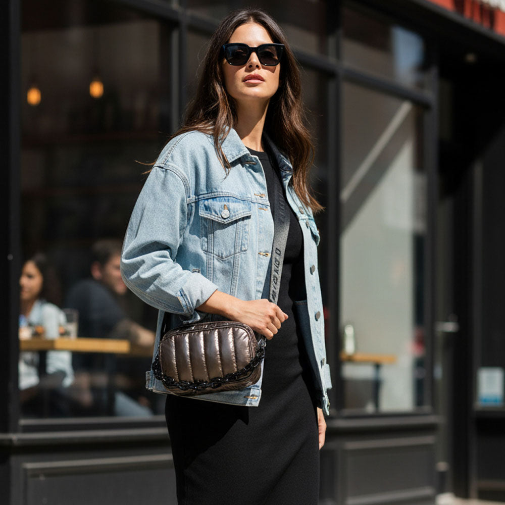 Woman wearing a denim jacket and sunglasses, holding a bronze metallic handbag, standing in an urban setting.