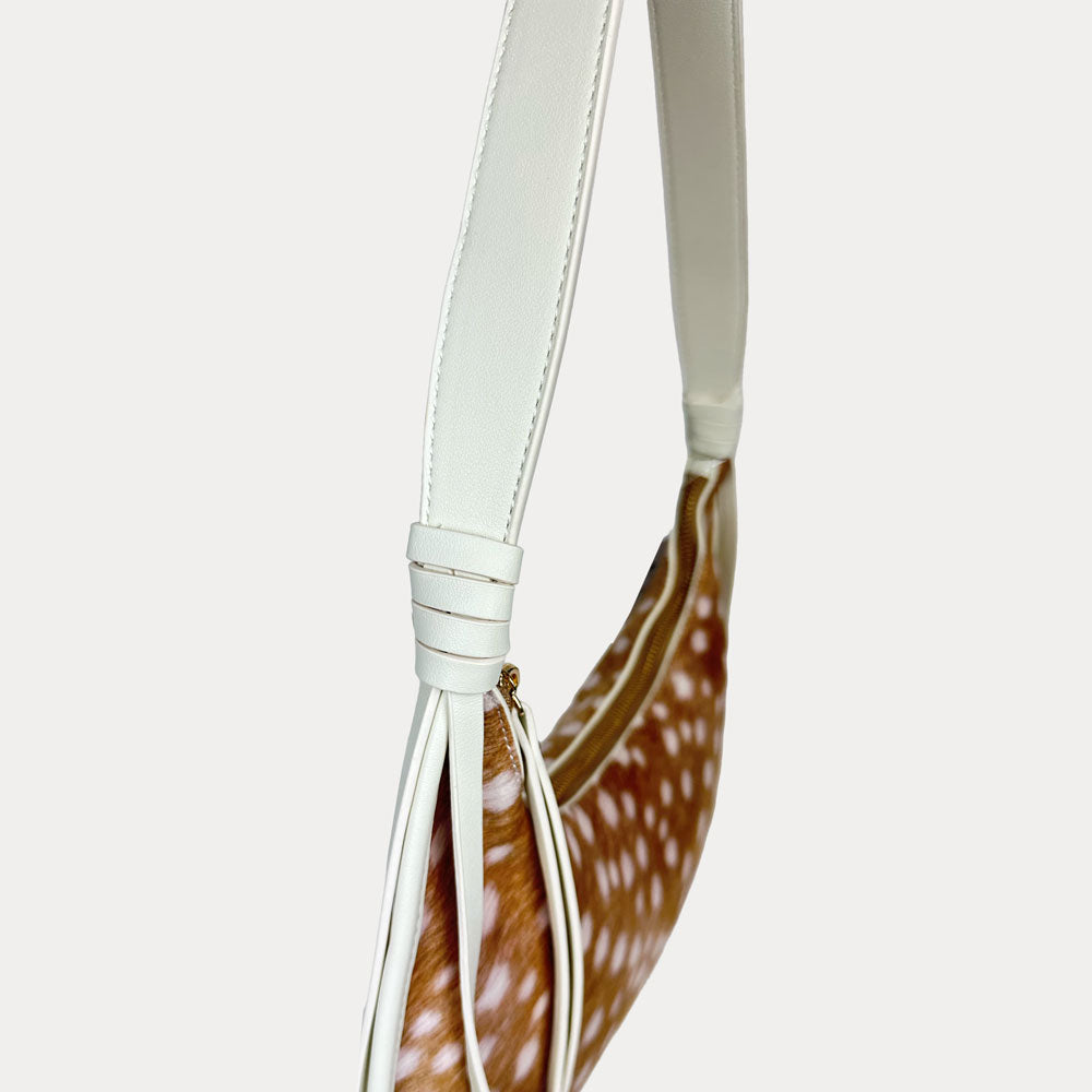 A close-up of a shoulder bag featuring a brown and white deer animal print body with a cream vegan leather strap and tassel detail, set against a solid light gray background.