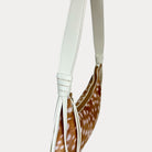A close-up of a shoulder bag featuring a brown and white deer animal print body with a cream vegan leather strap and tassel detail, set against a solid light gray background.