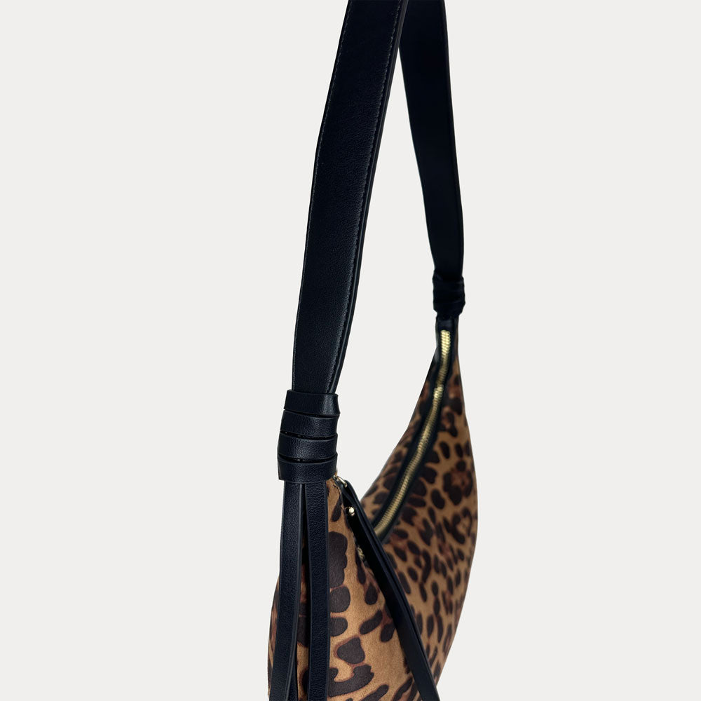 A close-up of a shoulder bag featuring a brown and black leopard print pattern with a black vegan leather strap set against a solid light gray background.