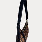 A close-up of a shoulder bag featuring a brown and black leopard print pattern with a black vegan leather strap set against a solid light gray background.