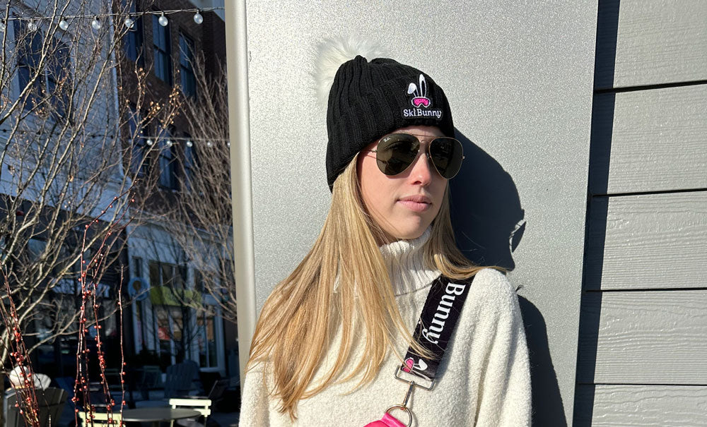 Person wearing a black beanie and sunglasses, standing against a gray wall with a blurred outdoor background.