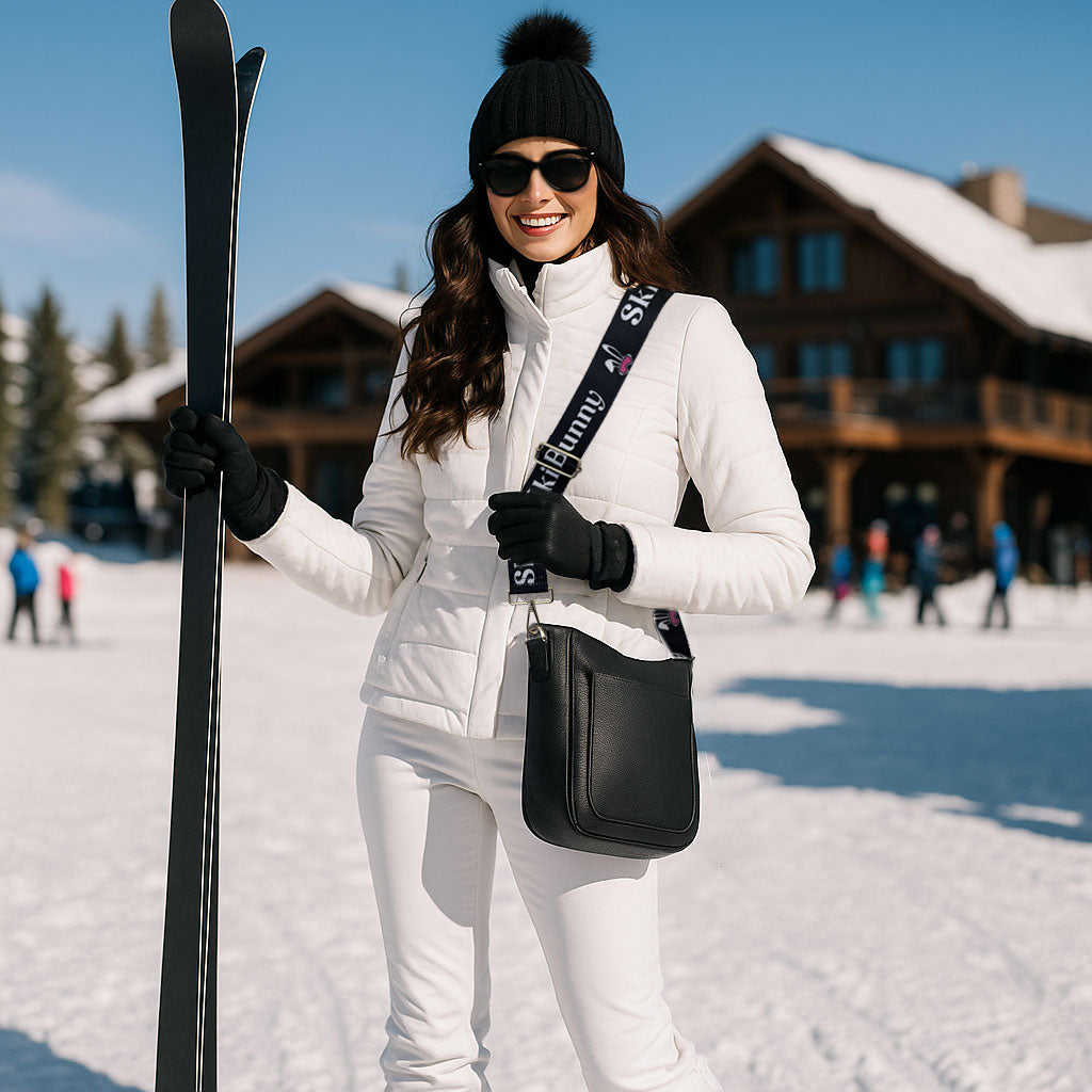 Woman in ski attire holding skis and a black bag in a snowy landscape with a ski lodge.