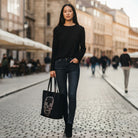 Woman in black outfit holding a black tote bag with a face design on a city street.