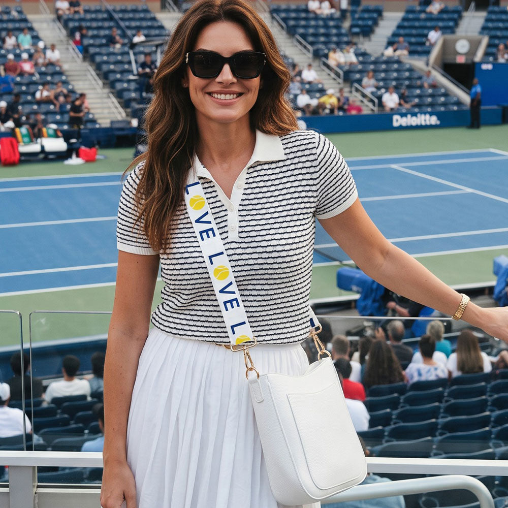 A woman stands in the standard a tennis match wearing a navy and white striped short-sleeve polo shirt, a white pleated skirt, black sunglasses, and a white crossbody bag with white 'LOVE' strap. The background shows stadium seating and a blue tennis court.