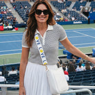 A woman stands in the standard a tennis match wearing a navy and white striped short-sleeve polo shirt, a white pleated skirt, black sunglasses, and a white crossbody bag with white 'LOVE' strap. The background shows stadium seating and a blue tennis court.
