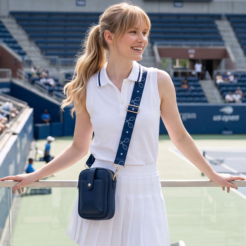 A woman on a at a tennis match in the stands a white sleeveless polo shirt and white skirt, carrying a navy blue crossbody bag with a navy bag strap featuring white tennis racket motifs. The background shows stadium seating and a tennis court.