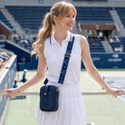 A woman on a at a tennis match in the stands a white sleeveless polo shirt and white skirt, carrying a navy blue crossbody bag with a navy bag strap featuring white tennis racket motifs. The background shows stadium seating and a tennis court.